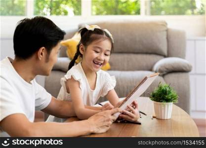 Asian girl reading tale story book with her father in living room while city lockdown from coronavirus covid-19 pandemic. Domestic life dad and daughter concept.