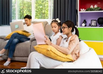 Asian girl reading tale story book with her mom in living room with dad work from home while city lockdown from coronavirus covid-19 pandemic. Domestic life family and work at home concept.