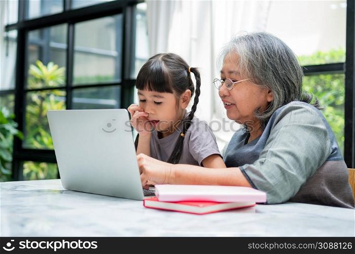 Asian Grandmother with her two grandchildren having fun and playing education games online with a computer notebook at home in the living room. Concept of online education and caring from parents.