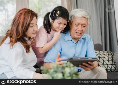 Asian grandparents and granddaughter using tablet at home. Senior Chinese, grandpa and grandma happy spend family time relax with young girl checking social media, lying on sofa in living room concept