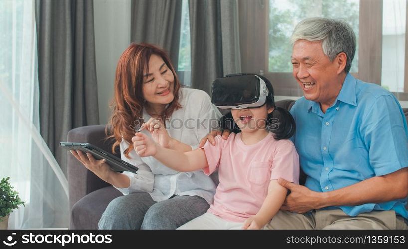 Asian grandparents and granddaughter using virtual reality and tablet play games at home. Senior Chinese, grandpa and grandma happy relax with young girl lying on sofa in living room concept.
