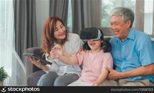 Asian grandparents and granddaughter using virtual reality and tablet play games at home. Senior Chinese, grandpa and grandma happy relax with young girl lying on sofa in living room concept.