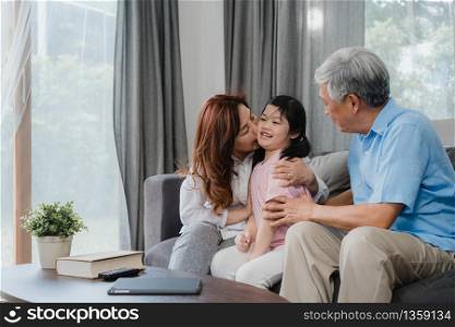 Asian grandparents kiss granddaughter cheek at home. Senior Chinese, old generation, grandfather and grandmother using family time relax with young girl kid lying on sofa in living room concept.