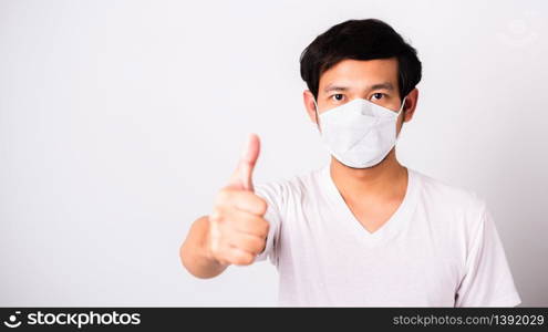 Asian handsome Man wearing surgical hygienic protective cloth face mask against coronavirus he show finger thumb for good, studio shot isolated white background, COVID-19 medical concept