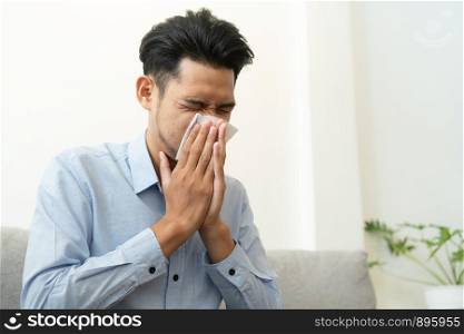 Asian handsome of man having flu season and sneeze using paper tissues sitting on sofa at home, Health and illness concepts