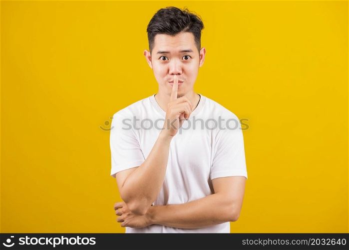 Asian handsome young man holding index finger on lips asking for silence or secret gesturing, studio shot isolated on yellow background, making shh shush gesture concept