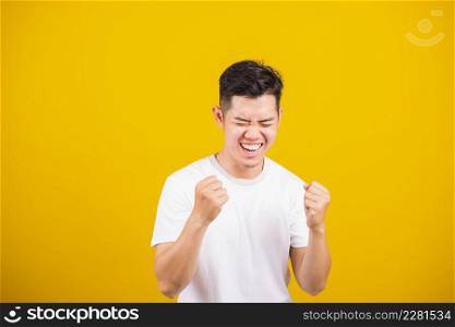Asian handsome young man smile positive shaking hands enthusiastic shouting yes for win competition, male raising his fists Yes! with smiling delighted face, studio shot isolated on yellow background