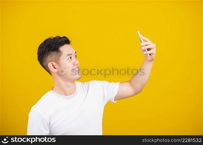 Asian handsome young man smiling positive saying hi talking by video call with mobile phone, excited happy male taking selfie on smartphone studio shot isolated on yellow background