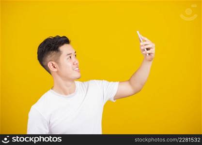 Asian handsome young man smiling positive saying hi talking by video call with mobile phone, excited happy male taking selfie on smartphone studio shot isolated on yellow background