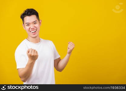 Asian handsome young man smiling positive shaking hands enthusiastic shouting yes for win competition, male raising his fists with smiling delighted face, studio shot isolated on yellow background