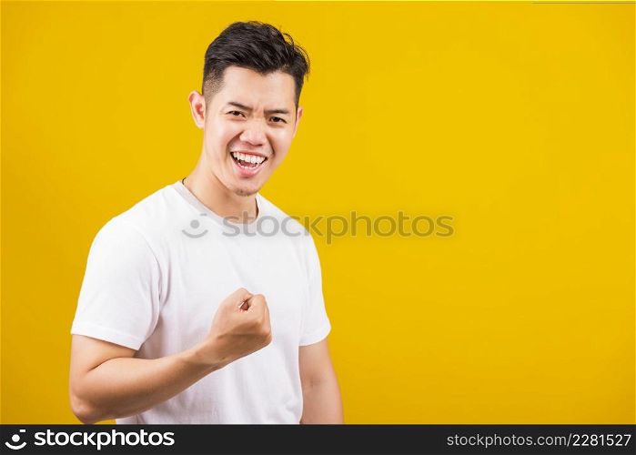 Asian handsome young man smiling positive shaking hands enthusiastic shouting yes for win competition, male raising his fists with smiling delighted face, studio shot isolated on yellow background