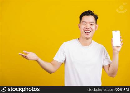 Asian handsome young man smiling positive showing mobile phone blank screen with another hand palm open empty space, excited happy male hold smartphone studio shot isolated on yellow background