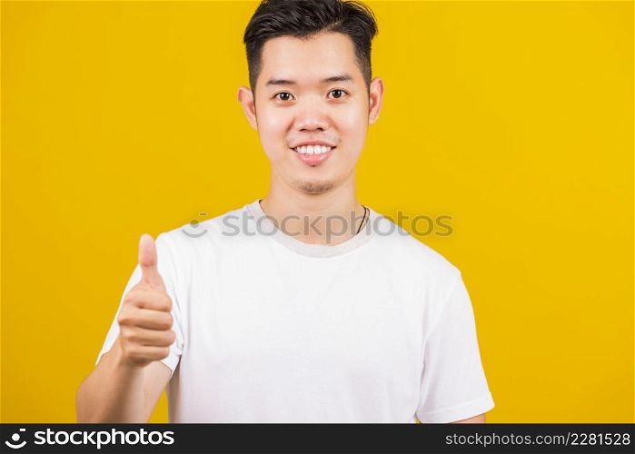 Asian handsome young man smiling positive showing thumbs up gesture good or like sign looking to camera, studio shot isolated on yellow background