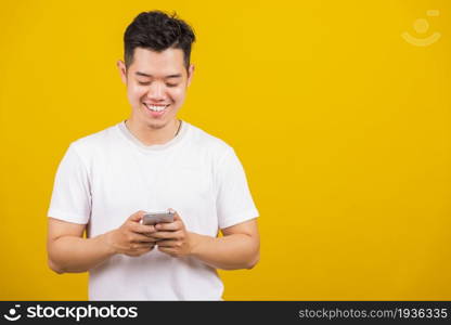 Asian handsome young man smiling positive using smartphone to get in touch screen or typing message, male reading good news message enjoy mobile application, studio shot isolated on yellow background