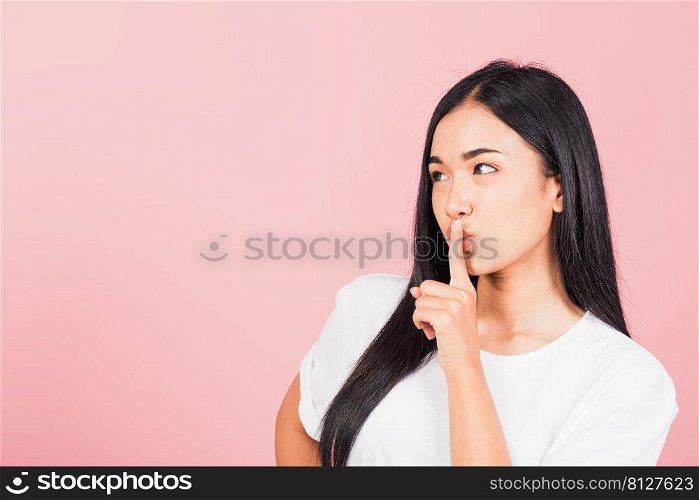 Asian happy portrait beautiful cute young woman smile stand making finger on lips mouth silent quiet gesture studio shot isolated on pink background, Thai female looking to side away with copy space