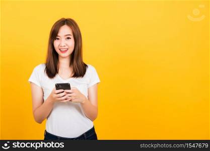 Asian happy portrait beautiful cute young woman teen smile standing playing game or writing message on smartphone looking to camera isolated, studio shot on yellow background with copy space