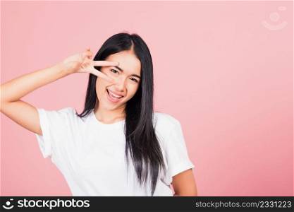 Asian happy portrait beautiful cute young woman teen smile standing showing finger making v-sign victory symbol near eye looking to camera studio shot isolated on pink background with copy space