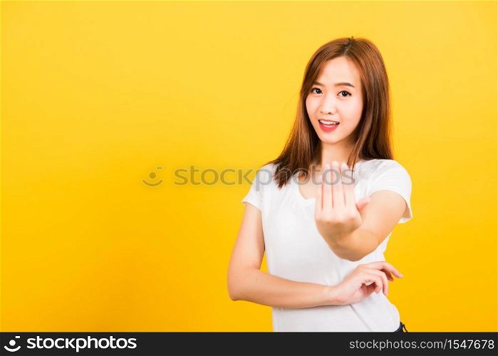 Asian happy portrait beautiful cute young woman teen smile standing wear white t-shirt making gesture hand inviting to come looking to camera isolated, studio shot on yellow background with copy space