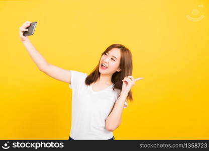 Asian happy portrait beautiful cute young woman teen smiling standing wear t-shirt mak selfie photo, video call on smartphone, point finger back isolated, studio shot yellow background with copy space