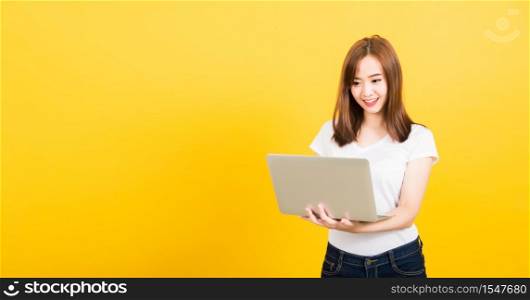 Asian happy portrait beautiful cute young woman teen smiling standing wear t-shirt using laptop computer and showing thumb up looking to camera isolated, studio shot yellow background with copy space