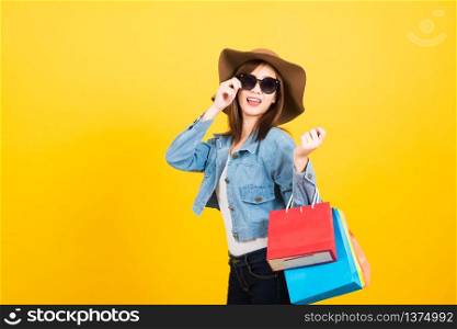 Asian happy portrait beautiful cute young woman teen smiling standing with sunglasses excited holding shopping bags multi color looking camera isolated, studio shot yellow background with copy space