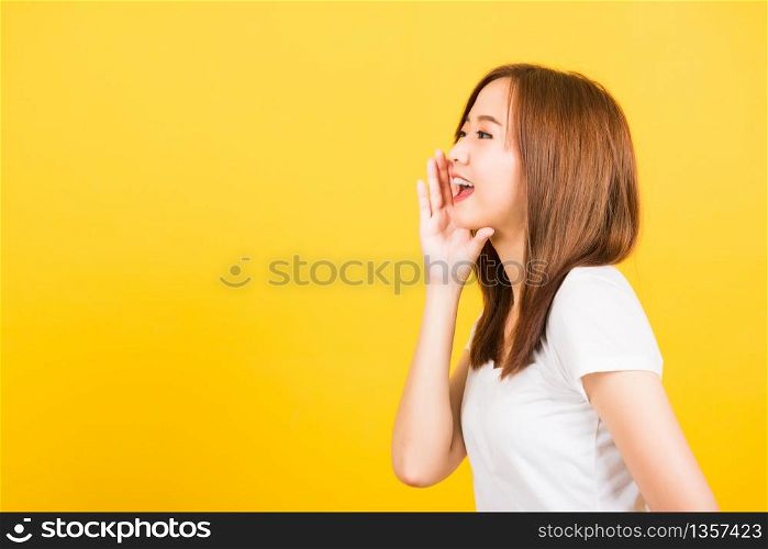 Asian happy portrait beautiful cute young woman teen standing wear t-shirt hand on mouth talking whispering secret rumor looking to side isolated, studio shot on yellow background with copy space