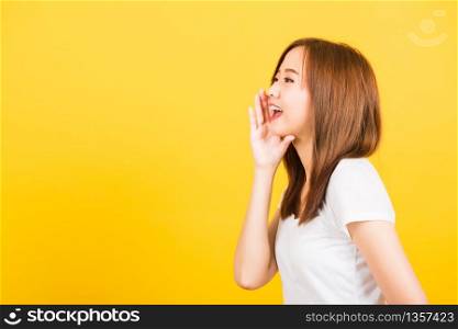 Asian happy portrait beautiful cute young woman teen standing wear t-shirt hand on mouth talking whispering secret rumor looking to side isolated, studio shot on yellow background with copy space