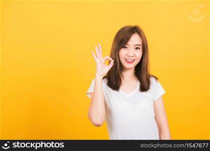 Asian happy portrait beautiful cute young woman teen standing wear t-shirt showing gesturing ok sign with fingers looking to camera isolated, studio shot on yellow background with copy space for text