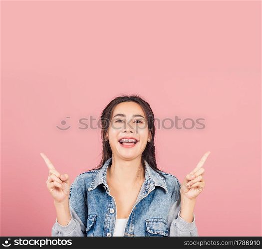 Asian happy portrait beautiful cute young woman wear denim standing makes gesture two fingers point upwards above presenting product something, studio shot isolated on pink background with copy space