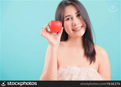 Asian happy portrait beautiful young woman standing smile holding red heart on hand near face on blue background with copy space for text