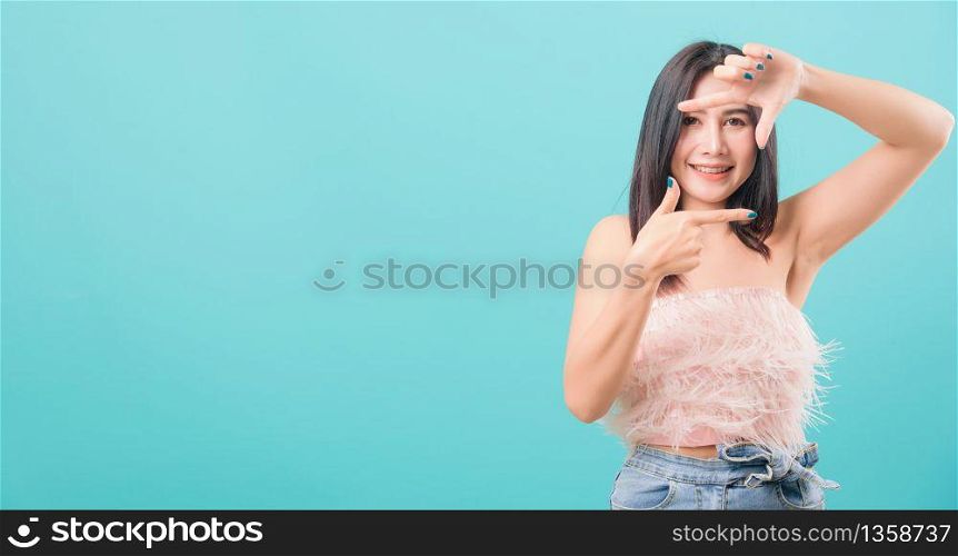 Asian happy portrait beautiful young woman standing smiling showing frame finger sign and looking to camera isolated on blue background with copy space for text