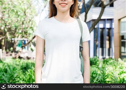 Asian hipster girl long brown hair in white blank t-shirt is standing in the middle of street. A female in street wear is standing on a green urban background. Empty mock up space for text or design.