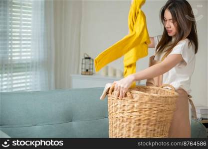 Asian housewife woman holding basket with heap of different clothes on sofa in living room, Beautiful housekeeper doing housework holds wooden basket of clean clothes to washing at home