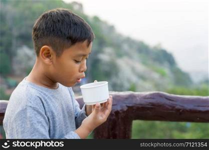 Asian kid boy is drinking water from the glass. With mountain views background