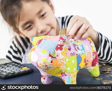 Asian kid is putting money in to a colorful piggy coin bank with calculator on wooden table