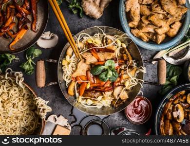 Asian kitchen table with food bowls, wok , stir fry , chopsticks and ingredients on background, top view