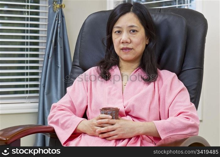 Asian lady holding cup of coffee in bathrobe while relaxing in massage chair at home