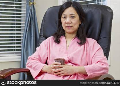 Asian lady holding cup of coffee in bathrobe while relaxing in massage chair at home