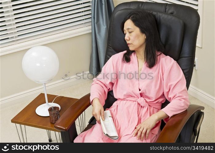 Asian lady in bathrobe holding magazine while resting in massage chair at home