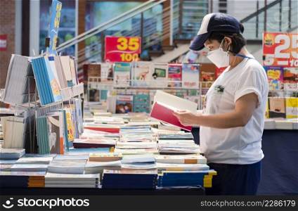 Asian LGBT woman in casual style with cap and protective mask choosing books at bookstore in shopping mall area
