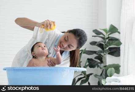 Asian little baby bathing in bathtub and looking at sponge which young mother squeeze water out while mom shower her cute daughter at home, Baby bathing at home concept. White background