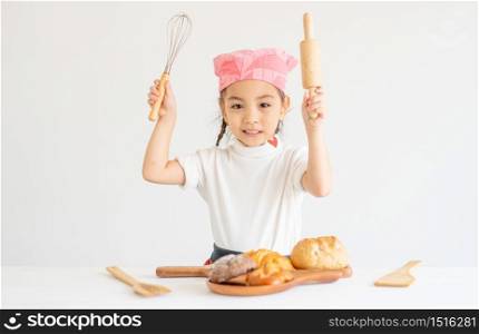 Asian little chef girl hold rolling pin and egg whisk stand in front of bread with white background.