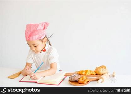 Asian little chef girl prepare and record her recipes about bread and cake cooking on notebook with white background.