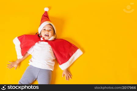 Asian little cute child girl smile, Top view of a kid dressed in red Santa Claus hat the concept of holiday Christmas Xmas day or Happy new year, isolated lying on yellow background