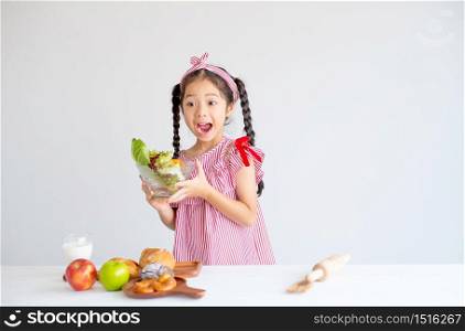 Asian little cute girl show action of surprise and hold bowl of vegetables with the apples and bread on table.