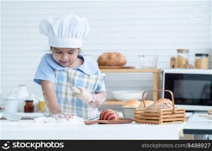Asian little cute kid girl wear chef hat and apron with flour mess up on face kneading dough bread by hand and rolling pin in kitchen at home or cooking class at school. Child education concept