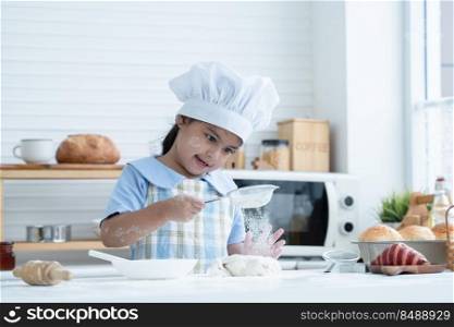 Asian little cute kid girl wear chef hat and apron with flour mess up on face have fun sifting flour on dough bread in kitchen at home or baking class at school. Child education concept