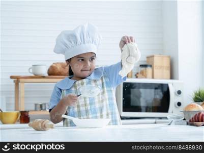 Asian little cute kid girl wear chef hat and apron with flour mess up on face have fun sifting flour and knead dough bread in kitchen at home or baking class at school. Child education concept