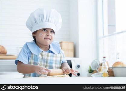 Asian little cute kid girl wear chef hat and apron with flour mess up on face holding wooden rolling pin kneading dough bread in kitchen at home or cooking class at school. Child education concept