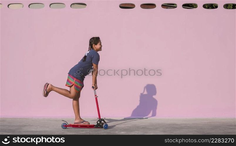 Asian little dark skinned girl has fun by riding kick scooter near pink wall on the patio outside of home